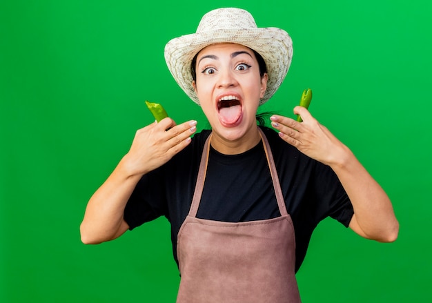 Jardinero de mujer hermosa joven en delantal y sombrero sosteniendo ají verde mirando al frente emocionado sacando la lengua de pie sobre la pared verde