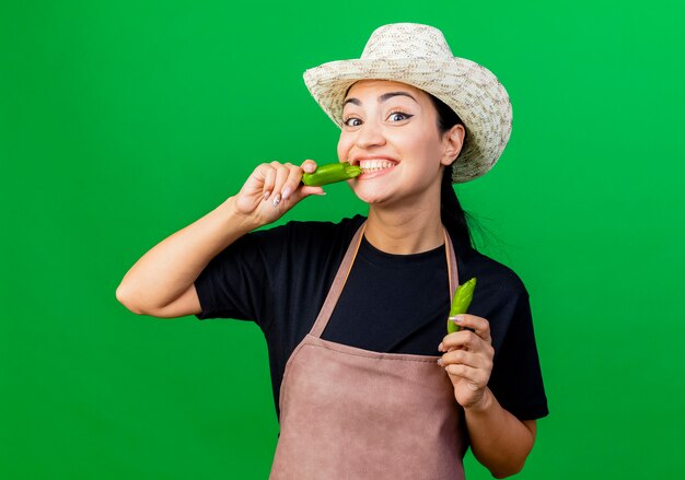 Jardinero de mujer hermosa joven en delantal y sombrero sosteniendo ají verde roto va a morderlo