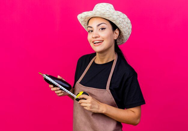 Jardinero de mujer hermosa joven en delantal y sombrero sosteniendo berenjena y cinta métrica mirando al frente sonriendo con cara feliz de pie sobre la pared rosa