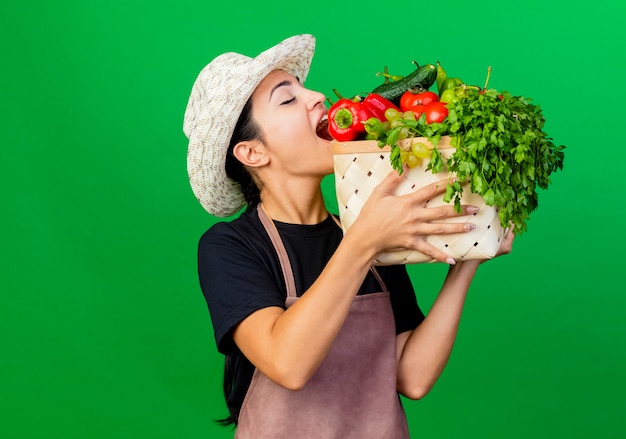 Jardinero de mujer hermosa joven en delantal y sombrero sosteniendo cajón lleno de verduras mordiendo pimiento rojo