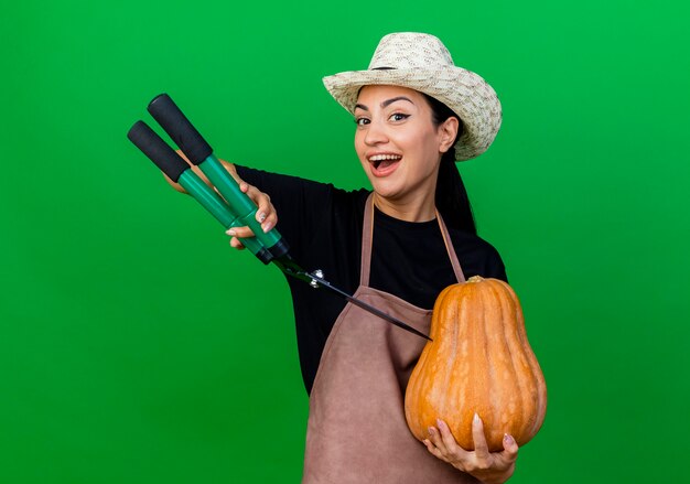 Foto gratuita jardinero de mujer hermosa joven en delantal y sombrero sosteniendo calabaza y cortasetos mirando al frente sonriendo con cara feliz de pie sobre la pared verde