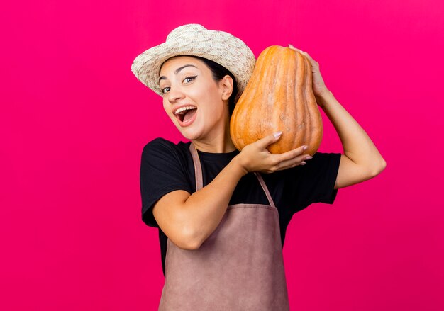 Jardinero de mujer hermosa joven en delantal y sombrero sosteniendo calabaza mirando al frente feliz y emocionado de pie sobre la pared rosa