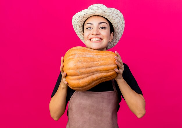 Jardinero de mujer hermosa joven en delantal y sombrero sosteniendo calabaza mirando al frente sonriendo alegremente de pie sobre la pared rosa