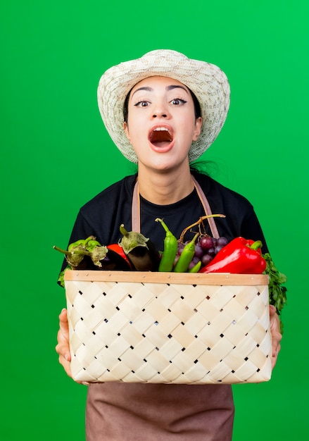 Jardinero de mujer hermosa joven en delantal y sombrero sosteniendo una canasta llena de verduras gritando con cara feliz de pie sobre la pared verde