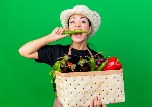 Jardinero de mujer hermosa joven en delantal y sombrero sosteniendo la canasta llena de verduras mordiendo ají verde de pie sobre la pared verde