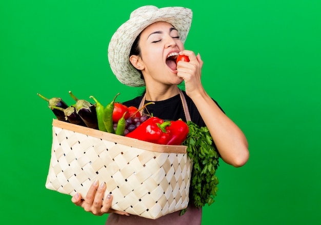 Jardinero de mujer hermosa joven en delantal y sombrero sosteniendo la cesta llena de verduras mordiendo tomate de pie sobre la pared verde