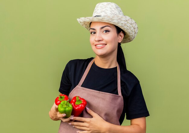 Jardinero de mujer hermosa joven en delantal y sombrero sosteniendo coloridos pimientos mirando al frente sonriendo alegremente de pie sobre la pared verde claro