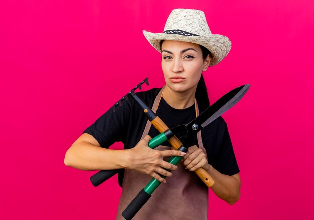 Jardinero de mujer hermosa joven en delantal y sombrero sosteniendo cortasetos y mini rastrillo mirando al frente con cara seria de pie sobre la pared rosa