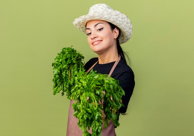 Jardinero de mujer hermosa joven en delantal y sombrero sosteniendo hierbas frescas mirando al frente sonriendo alegremente de pie sobre la pared verde claro