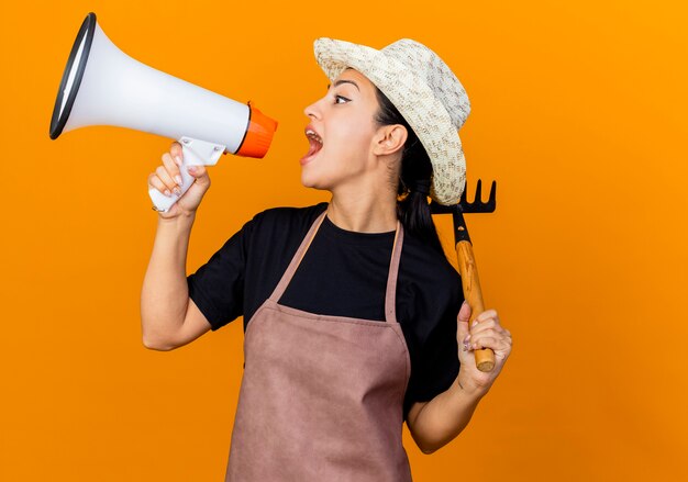 Jardinero de mujer hermosa joven en delantal y sombrero sosteniendo mini rastrillo gritando al megáfono de pie sobre la pared naranja