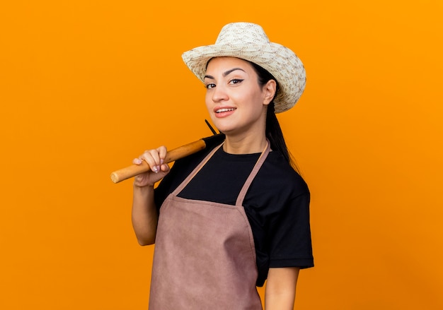 Jardinero de mujer hermosa joven en delantal y sombrero sosteniendo mini rastrillo mirando al frente sonriendo con cara feliz de pie sobre la pared naranja