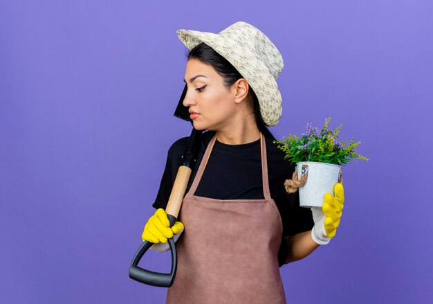 Jardinero de mujer hermosa joven en delantal y sombrero sosteniendo una pala y una planta en maceta mirando confiado de pie sobre la pared azul