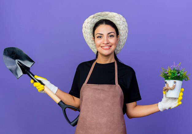 Jardinero de mujer hermosa joven en delantal y sombrero sosteniendo pala y planta en maceta sonriendo alegremente de pie sobre la pared azul