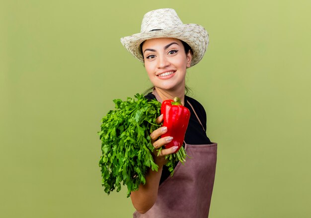 Jardinero de mujer hermosa joven en delantal y sombrero sosteniendo pimiento rojo y hierbas frescas sonriendo alegremente de pie sobre la pared verde claro