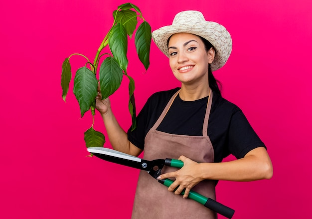 Jardinero de mujer hermosa joven en delantal y sombrero sosteniendo planta y cortasetos mirando al frente sonriendo alegremente de pie sobre la pared rosa