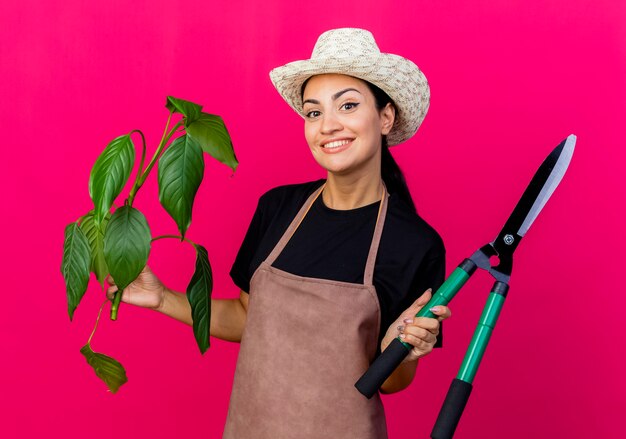 Jardinero de mujer hermosa joven en delantal y sombrero sosteniendo planta y cortasetos sonriendo con cara feliz