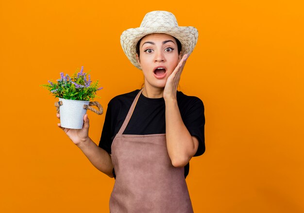 Jardinero de mujer hermosa joven en delantal y sombrero sosteniendo planta en maceta mirando al frente siendo sorprendido y asombrado de pie sobre la pared naranja