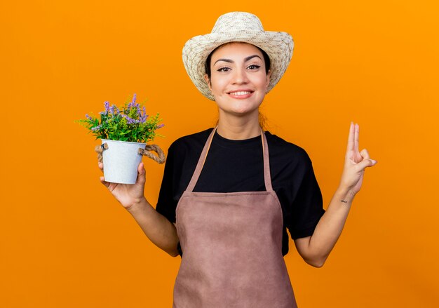 Jardinero de mujer hermosa joven en delantal y sombrero sosteniendo planta en maceta mirando al frente sonriendo seguro mostrando el número dos de pie sobre la pared naranja