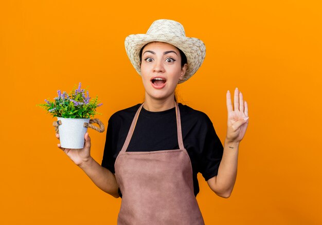 Jardinero de mujer hermosa joven en delantal y sombrero sosteniendo planta en maceta mirando al frente sorprendido mostrando el número cuatro de pie sobre la pared naranja