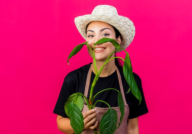 Jardinero de mujer hermosa joven en delantal y sombrero sosteniendo la planta mirando sonriendo con cara feliz de pie