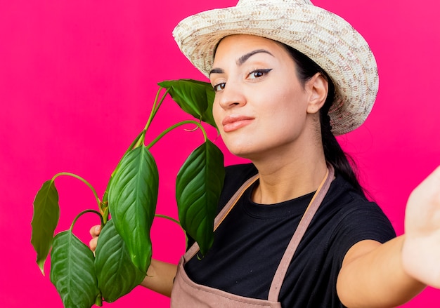 Foto gratuita jardinero de mujer hermosa joven en delantal y sombrero sosteniendo la planta sonriendo con cara feliz