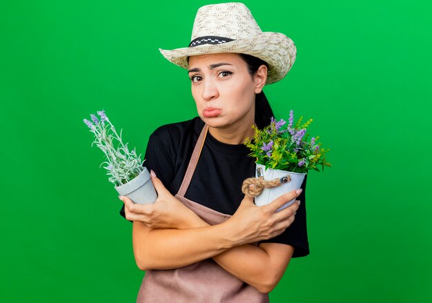 Foto gratuita jardinero de mujer hermosa joven en delantal y sombrero sosteniendo plantas en macetas con expresión triste