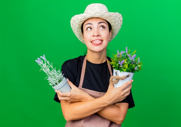 Jardinero de mujer hermosa joven en delantal y sombrero sosteniendo plantas en macetas mirando hacia arriba sacando la lengua de pie sobre la pared verde