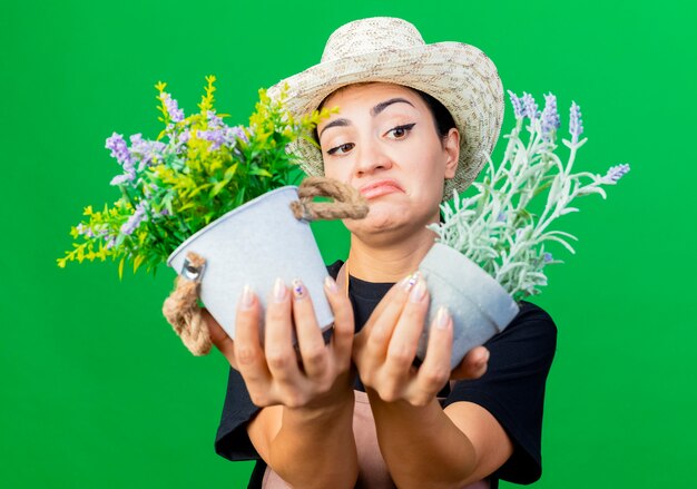 Jardinero de mujer hermosa joven en delantal y sombrero sosteniendo plantas en macetas mirando confundido y disgustado de pie sobre la pared verde