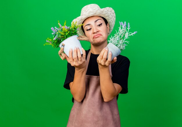 Jardinero de mujer hermosa joven en delantal y sombrero sosteniendo plantas en macetas mirando confundido y disgustado