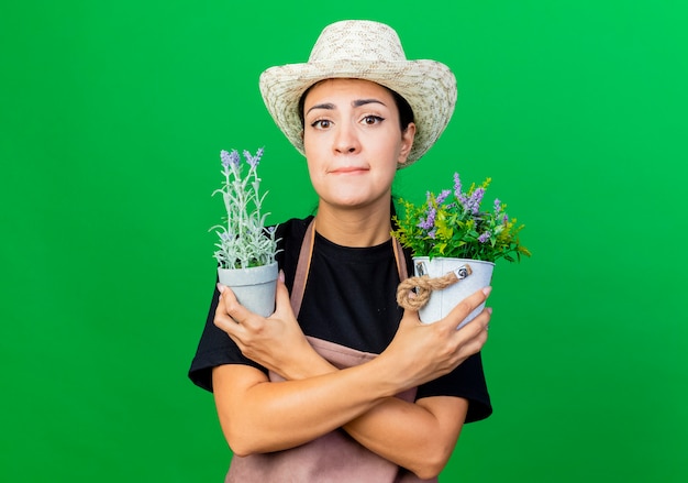 Jardinero de mujer hermosa joven en delantal y sombrero sosteniendo plantas en macetas preocupado