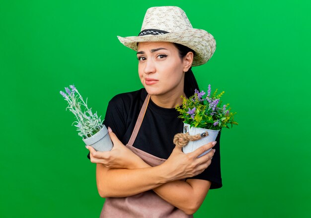 Jardinero de mujer hermosa joven en delantal y sombrero sosteniendo plantas en macetas sonriendo confundido