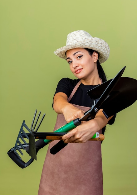 Foto gratuita jardinero de mujer hermosa joven en delantal y sombrero sosteniendo podadoras de setos y azadón mirando al frente sonriendo de pie sobre la pared verde claro