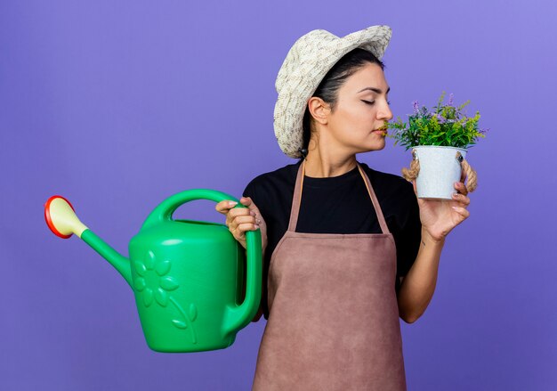 Jardinero de mujer hermosa joven en delantal y sombrero sosteniendo regadera mirando pland en macetas en otra mano de pie sobre la pared azul