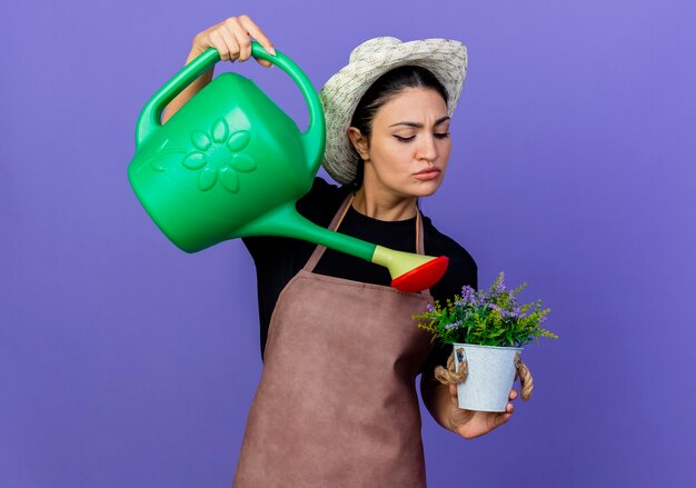 Jardinero de mujer hermosa joven en delantal y sombrero sosteniendo regadera, planta en maceta de riego de pie sobre la pared azul