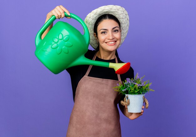 Jardinero de mujer hermosa joven en delantal y sombrero sosteniendo regadera, regando planta en maceta sonriendo alegremente de pie sobre la pared azul