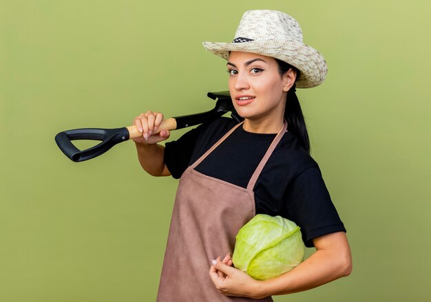 Jardinero de mujer hermosa joven en delantal y sombrero sosteniendo repollo y pala mirando al frente sonriendo de pie sobre la pared verde claro