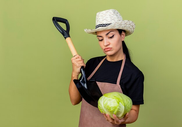 Jardinero de mujer hermosa joven en delantal y sombrero sosteniendo repollo y pala mirando repollo con expresión escéptica de pie sobre la pared verde claro