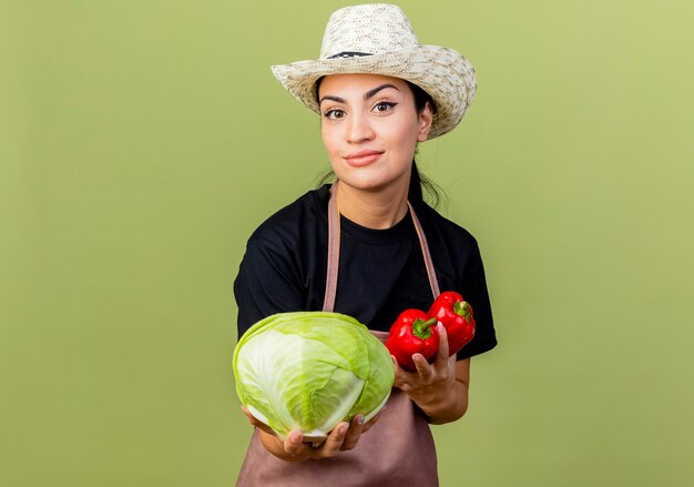 Jardinero de mujer hermosa joven en delantal y sombrero sosteniendo repollo y pimientos rojos sonriendo de pie sobre la pared verde claro