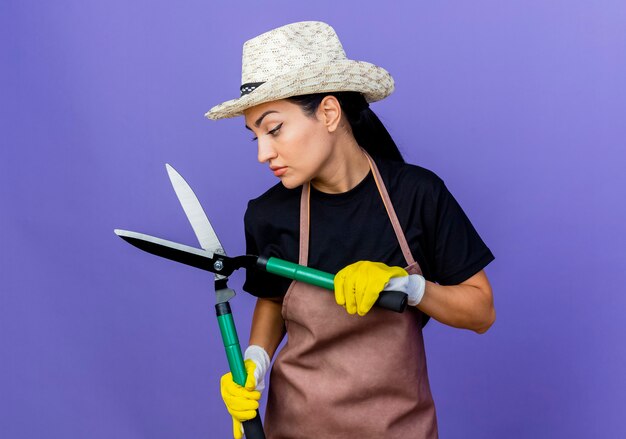 Jardinero de mujer hermosa joven en delantal y sombrero sosteniendo tijeras de podar buscando confianza de pie sobre la pared azul