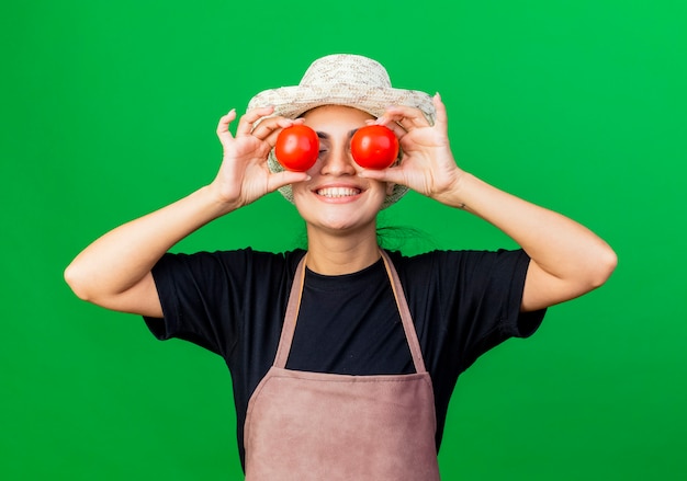 Jardinero de mujer hermosa joven en delantal y sombrero sosteniendo tomates cubriéndose los ojos con ellos sonriendo