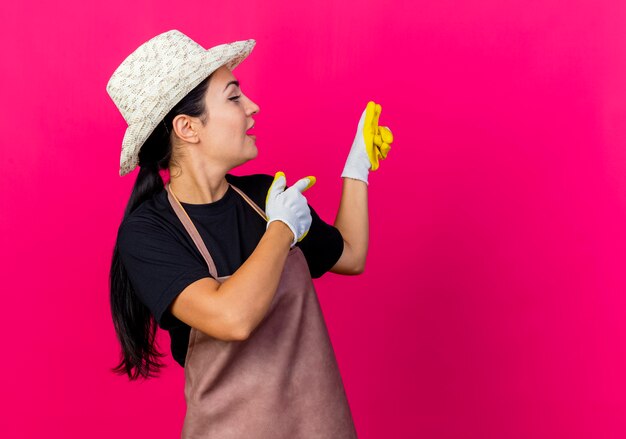 Jardinero de mujer hermosa joven en guantes de goma delantal y sombrero apuntando con el dedo índice hacia atrás siendo sorprendido de pie