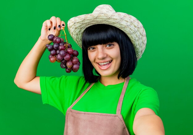 Jardinero mujer joven alegre en uniforme vistiendo sombrero de jardinería tiene uvas aisladas