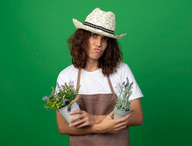 Jardinero de mujer joven disgustado en uniforme con sombrero de jardinería sosteniendo y cruzando flores en macetas aisladas en verde