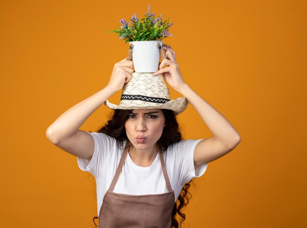 Jardinero mujer joven disgustado en uniforme con sombrero de jardinería sostiene maceta sobre la cabeza aislada en la pared naranja con espacio de copia