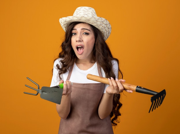 Jardinero mujer joven molesto en uniforme con sombrero de jardinería sostiene rastrillo y rastrillo azada aislado en la pared naranja con espacio de copia