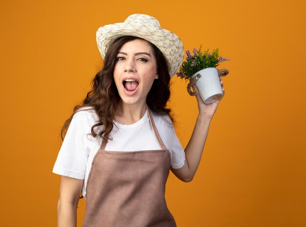 Jardinero mujer caucásica bastante alegre con sombrero y guantes de  jardinería sosteniendo maceta y mirando a pala aislado en la pared naranja  con espacio de copia