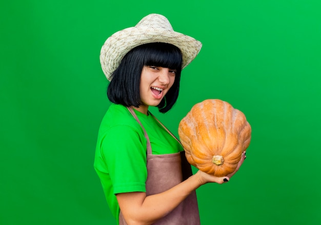 Jardinero mujer joven molesto en uniforme vistiendo sombrero de jardinería sosteniendo calabaza