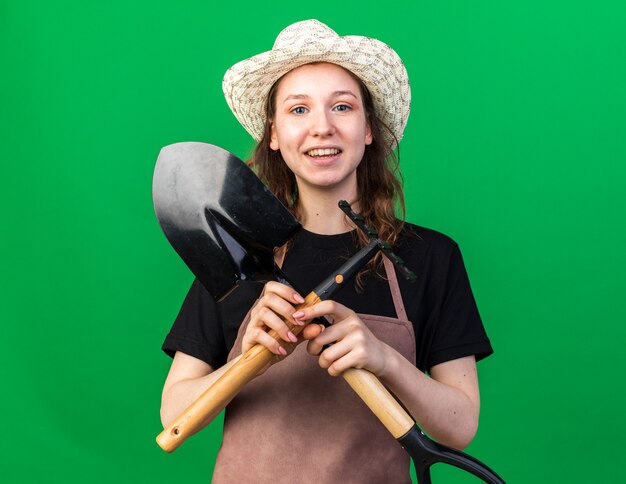 Jardinero mujer joven sonriente con sombrero de jardinería sosteniendo y cruzando la pala con rastrillo