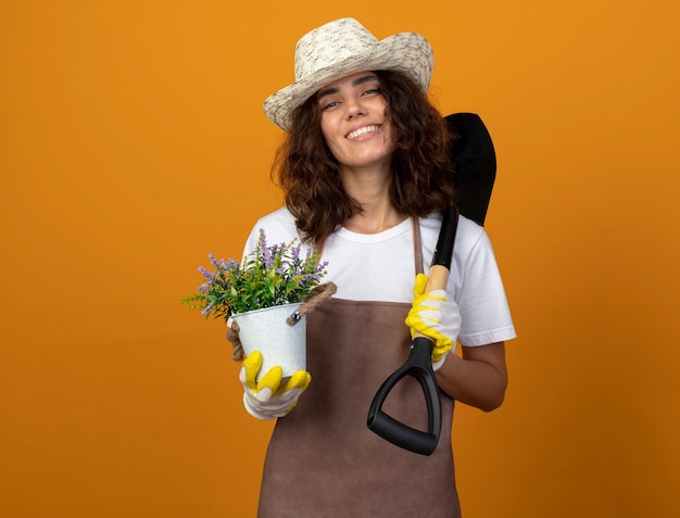 Foto gratuita jardinero de mujer joven sonriente en uniforme con sombrero y guantes de jardinería sosteniendo flores en maceta y poniendo pala en el hombro aislado en naranja