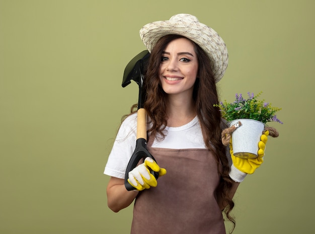 Jardinero mujer joven sonriente en uniforme con sombrero y guantes de jardinería sostiene maceta y pala en el hombro aislado en la pared verde oliva con espacio de copia